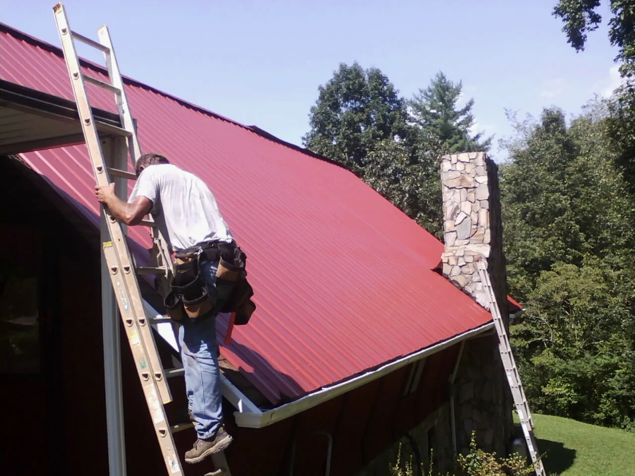 Roofer on a ladder beside a red metal roof and stone chimney — Soffit & Fascia Repair by Roofing Brothers in Crawford